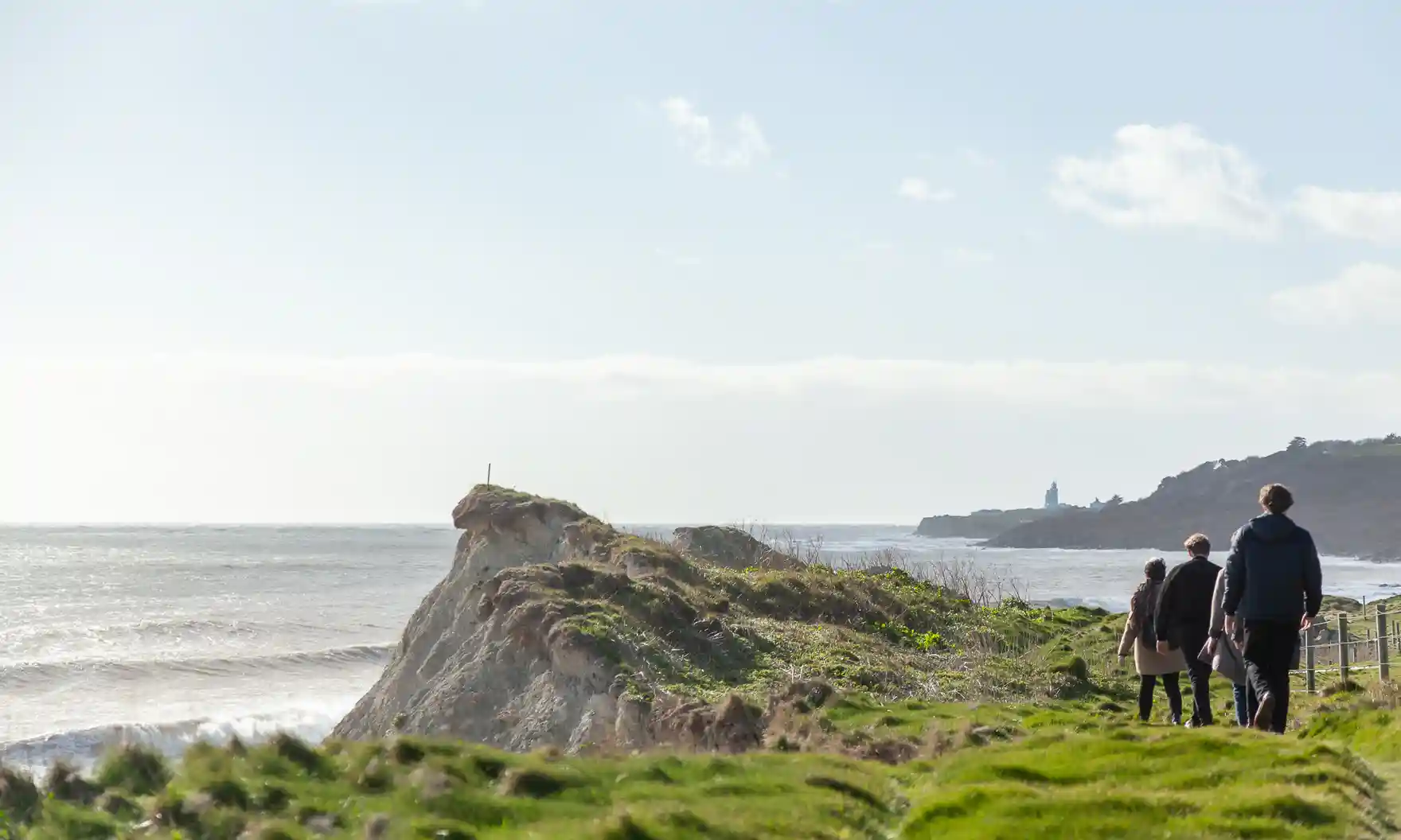 Image shows a clifftop walk with crashing waves on the coast below, people walking away from the camera toward St Catherine's Light House on the Isle of Wight. A perfect Winter wander.