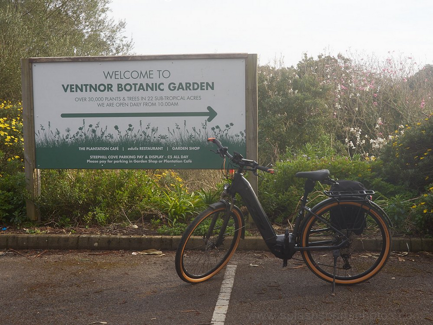 A electric bike stands in front of the Ventnor Botanic Garden sign with the blossom of a Magnolia tree behind.