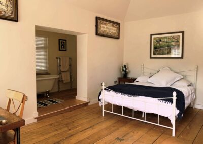 A view of a bedroom within one of the Ventnor Botanic Garden, self-catering cottages. Showing beautiful wooden floors, a metal bed with thick duvet and a view through to the bathroom with rolltop bath.