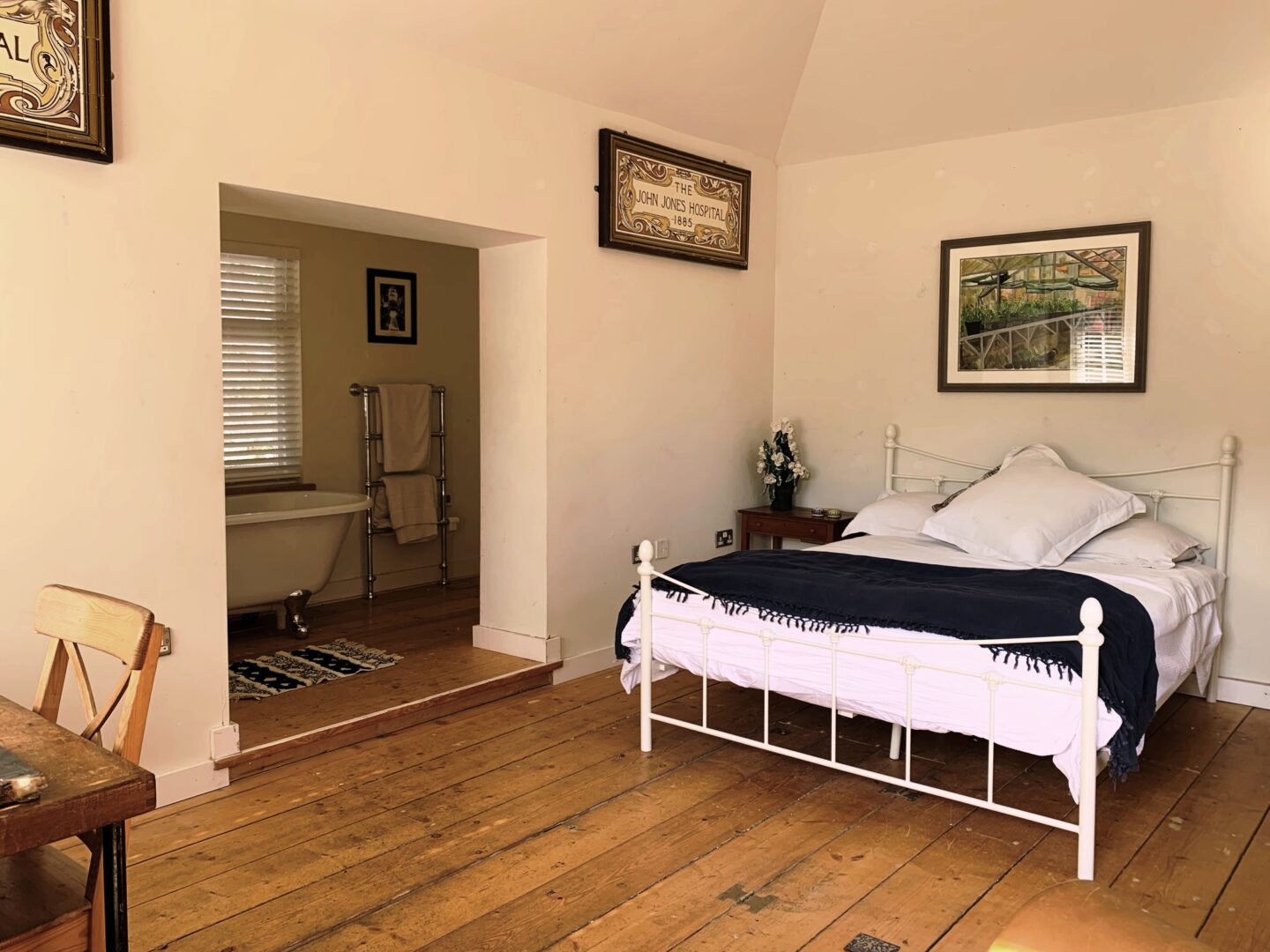 A view of a bedroom within one of the Ventnor Botanic Garden, self-catering cottages. Showing beautiful wooden floors, a metal bed with thick duvet and a view through to the bathroom with rolltop bath.